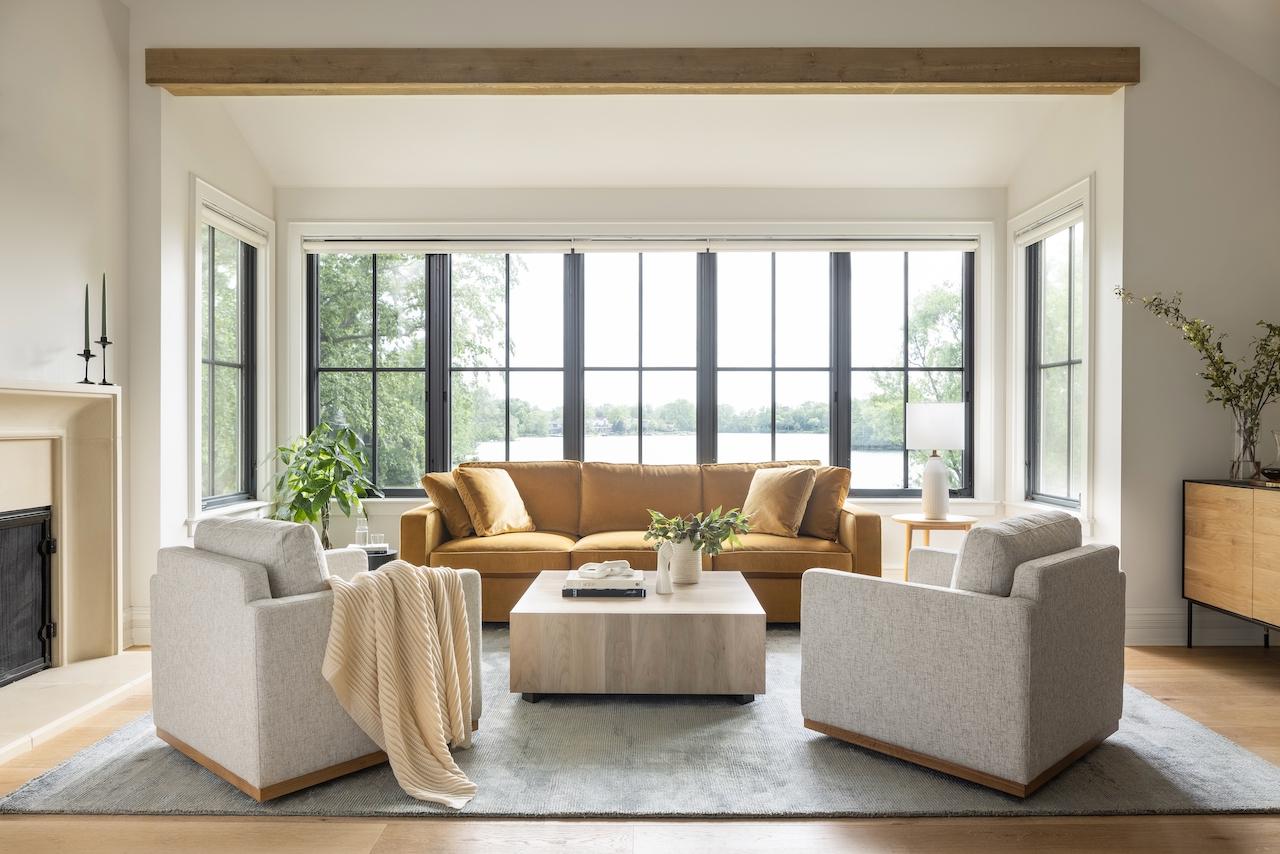 A contemporary living room highlights a mustard-yellow James 3 Seat Left Chaise 92" Sectional, accompanied by two gray armchairs and a light wood coffee table set upon a gray rug. Expansive windows reveal lush greenery outside, while a fireplace and potted plants enhance the warmth of the minimalist decor.