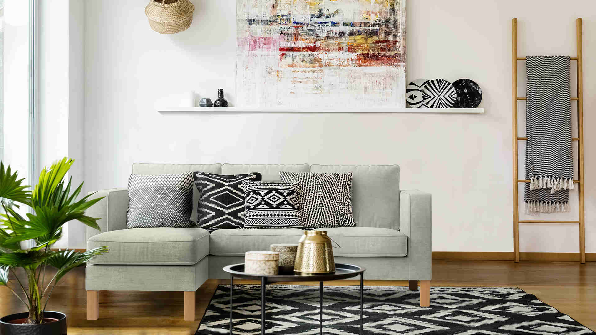 A modern living room with a light gray sectional sofa adorned with patterned cushions. A large abstract painting hangs above a white shelf decorated with vases. A ladder shelf holds blankets, and a potted plant sits beside the sofa. A black-and-white rug covers the floor.
