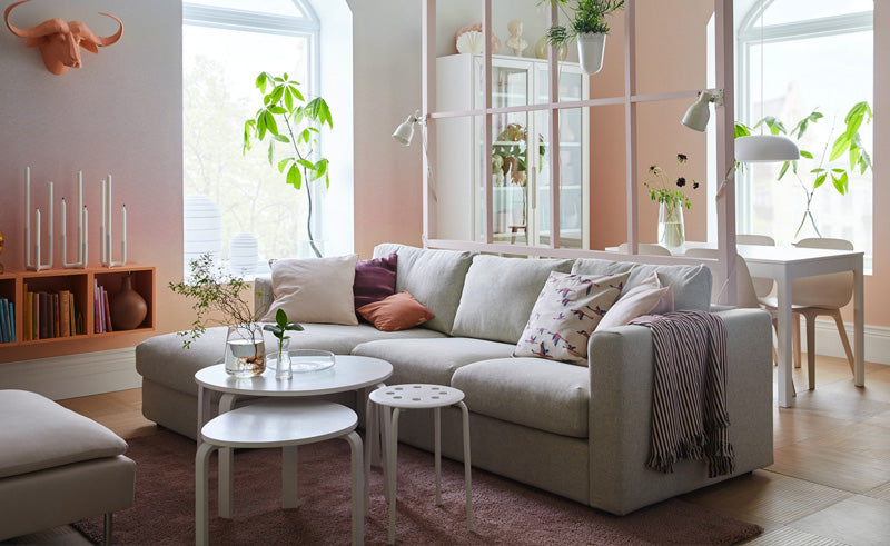 A cozy living room featuring a light gray sofa adorned with various pillows and a blanket. Nestled in front of the sofa are two round white coffee tables. Plants are placed throughout the space, enhancing the natural light streaming in from large windows.