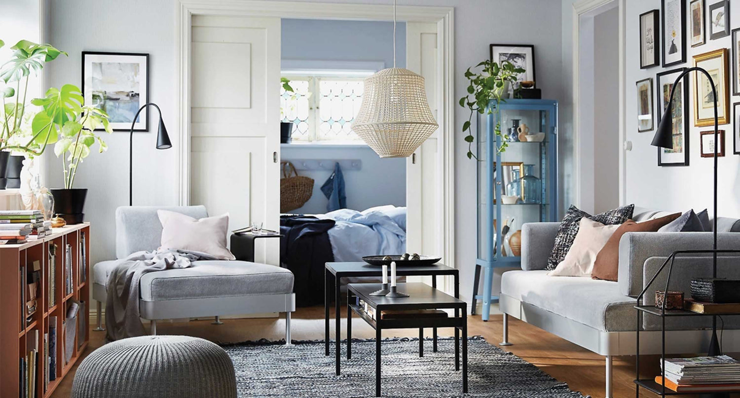 A cozy living room with gray sofas, a black coffee table, and a light woven pendant lamp. The space features plants, framed pictures, and a bookshelf. A bedroom with a neatly made bed is visible through sliding doors in the background.