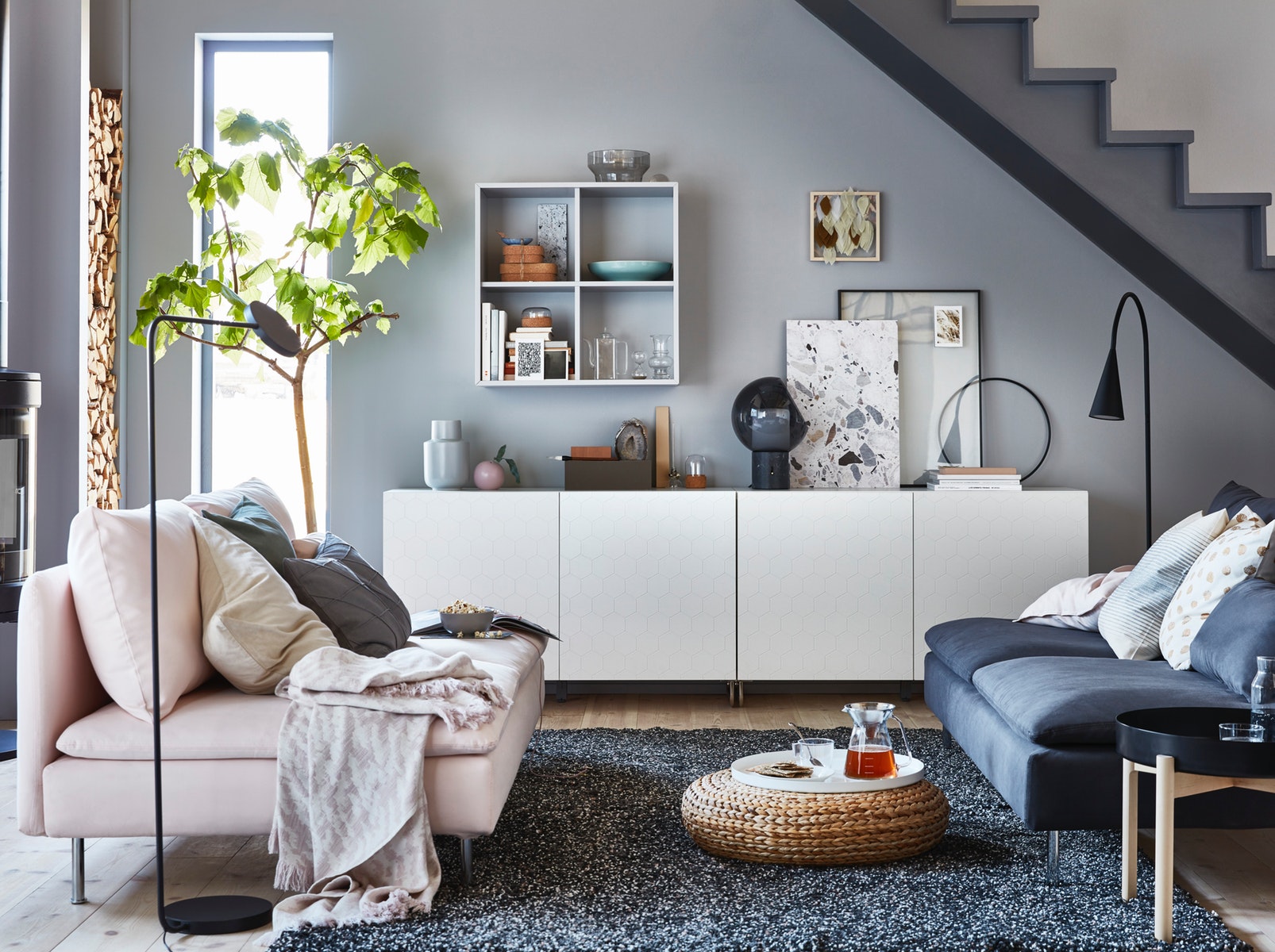 Modern living room with gray walls and light wood flooring. Features a dark gray sofa, light pink loveseat, potted plant, wall-mounted shelves with decor, white sideboard, and a black floor lamp. A woven ottoman and soft furnishings complete the cozy setup.