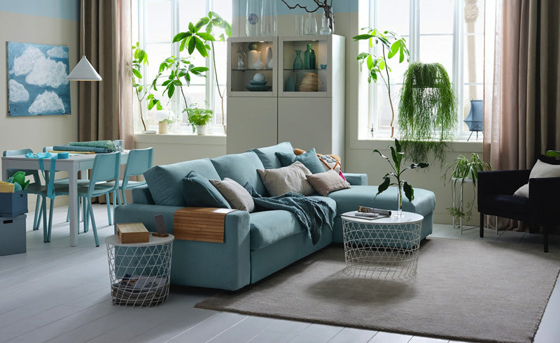 A modern, airy living room with a light blue sofa adorned with assorted cushions and throws. Indoor plants are thoughtfully placed around the room, and large windows allow ample natural light. A stylish wireframe coffee table sits on a beige rug in front of the sofa.