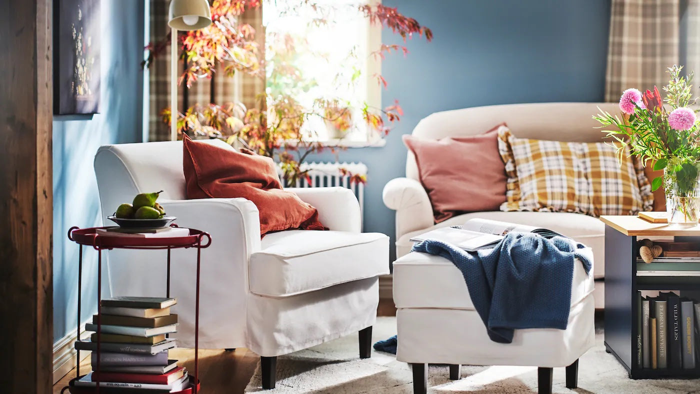 A cozy living room with a white armchair and ottoman, adorned with a rust-colored pillow and blue throw. A sofa with plaid and pink cushions sits by the window with autumn foliage outside. A side table holds a vase with colorful flowers. Stacks of books are on the floor.