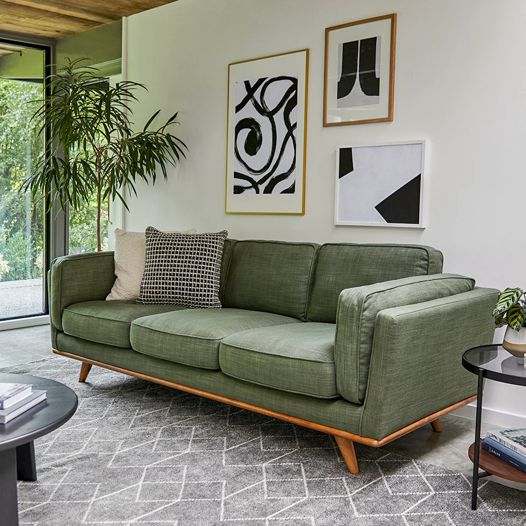 A modern living room showcases the Timber 90" Sofa adorned with patterned pillows. Above, abstract art pieces are displayed on the wall. A large plant is positioned nearby, and the room features a patterned rug alongside a round side table with decorative items.