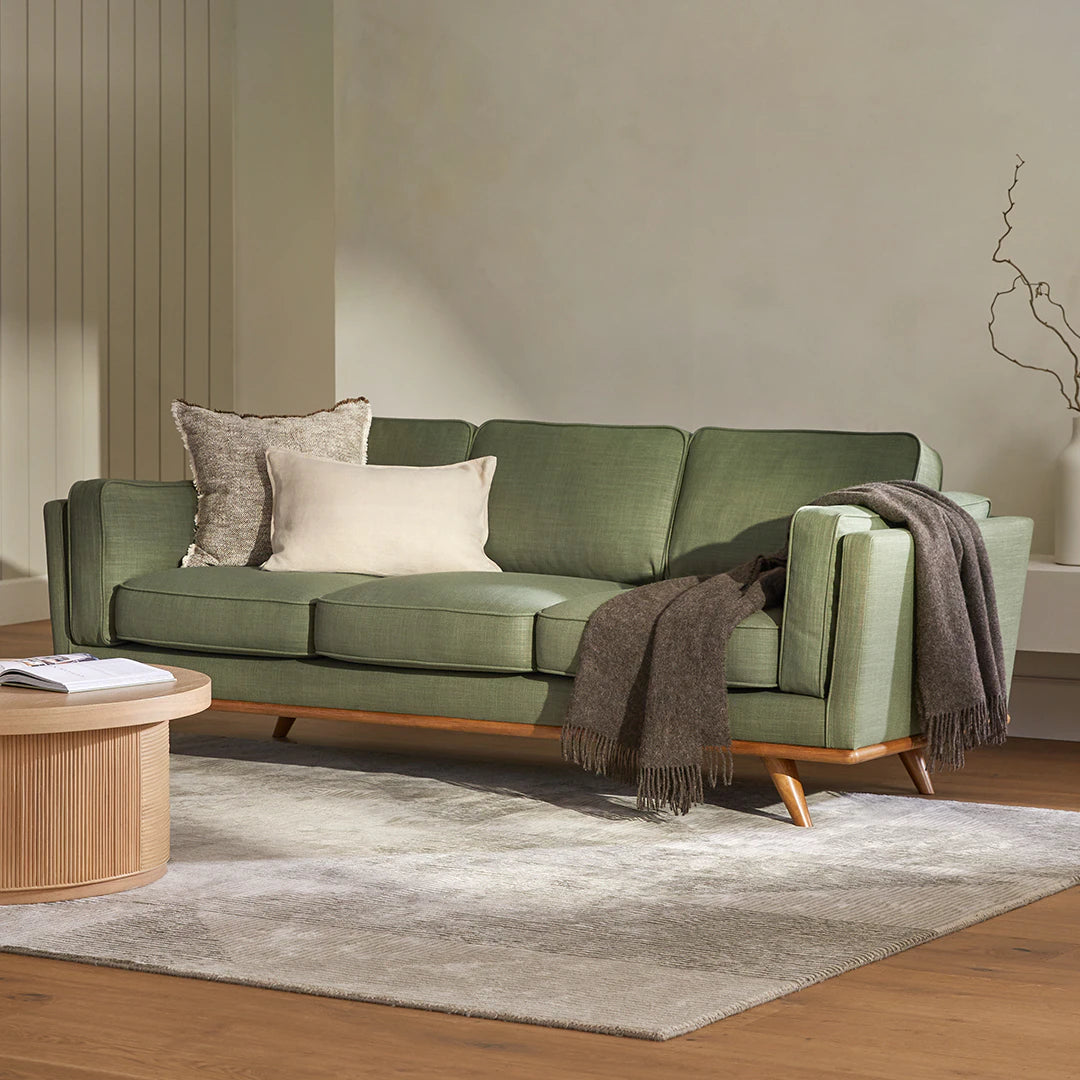 The Timber 90" Sofa, adorned with brown and beige cushions and a dark gray throw, stands on a light gray rug in a minimalist room. A round wooden coffee table is nearby, complementing the setting, while a decorative branch adds a touch of nature in the background.