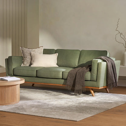 A modern living room showcases the Timber 90 Leather Sofa in Charme Tan, adorned with two cushions and a dark gray throw. The sofa is placed on a light gray area rug atop a wooden floor. A round wooden coffee table is nearby, and bare branches are displayed in a vase in the background.