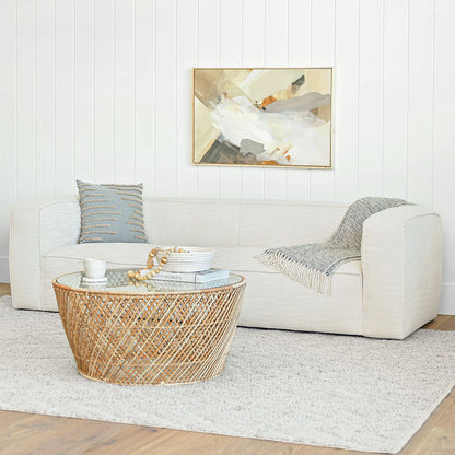 A modern living room features the Cigar 92" Leather Sofa in Rawhide Tan, adorned with a gray textured pillow and a draped blanket. A round wicker coffee table holds books and a beaded strand, while an abstract painting hangs on the white-paneled wall above a textured rug underfoot.