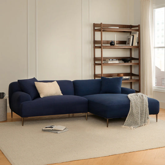 A cozy living room features the Abisko 94.5 Right Sectional in Aurora Blue, adorned with a cream pillow and a light gray throw blanket. A wooden shelving unit with decorative items rests against the wall, while soft natural light streams through a large window.