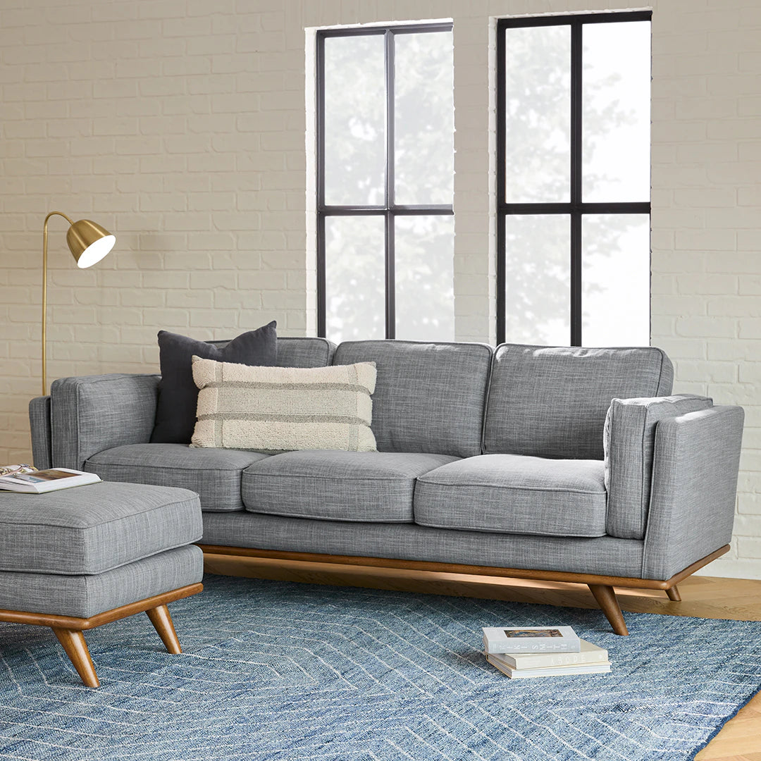 A modern living room featuring the Timber 90" sofa in gray fabric accompanied by a matching ottoman, all set elegantly on a blue patterned rug. The sofa is adorned with two cushions: one in solid gray and the other with stripes. A gold floor lamp stands beside it, while two tall windows allow natural light to flood the space.