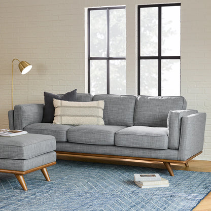 A modern living room with the Timber 90 Leather Sofa Charme Tan, embellished with throw pillows and an ottoman. It features a blue textured area rug and a floor lamp, while large windows offer a view of trees, allowing natural light to illuminate the light-colored brick wall.