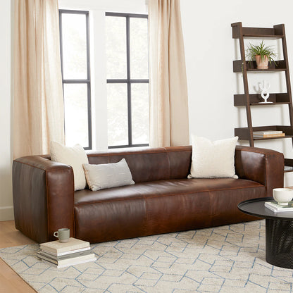 A cozy living room features a Cigar 92" Leather Sofa in Rawhide Tan, adorned with white and gray pillows. A stack of books and a mug are on the floor beside it. A ladder shelf with decor items stands nearby, and light streams through large windows with beige curtains.