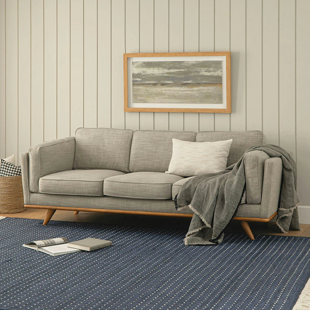 A cozy living room featuring a Timber 90 Leather Sofa in Charme Tan, adorned with a white pillow and a draped gray blanket. A seascape painting hangs above on beige paneled walls. On the floor lies a blue, dotted rug with a closed book and a notepad, while a wicker basket sits nearby.