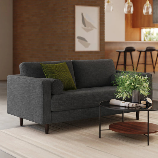 A modern living room showcasing a Sven 72 Tufted Loveseat Ratine Oxide adorned with green pillows. A sleek glass coffee table is set in front, featuring a potted plant and a magazine. The background reveals a brick wall adorned with abstract art and a kitchen area complete with barstools.