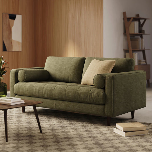 A contemporary living room showcases the Sven 72 Tufted Loveseat Ratine Oxide, offering a mid-century flair with cylindrical cushions and a beige throw pillow. A wooden coffee table adorned with a plant and books enhances the space, resting atop an eye-catching checker-patterned rug. In the background, a bookshelf and an abstract art piece complete the aesthetic.