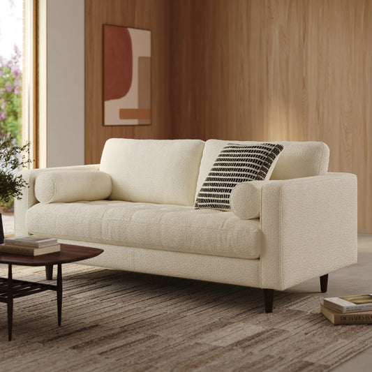 A contemporary living room showcases the Sven 72 Tufted Loveseat in Ratine Oxide, paired with a cylindrical pillow and a patterned cushion. A wooden coffee table supports a small plant. The room’s wooden walls are adorned with abstract art, while sunlight pours in through a large window.