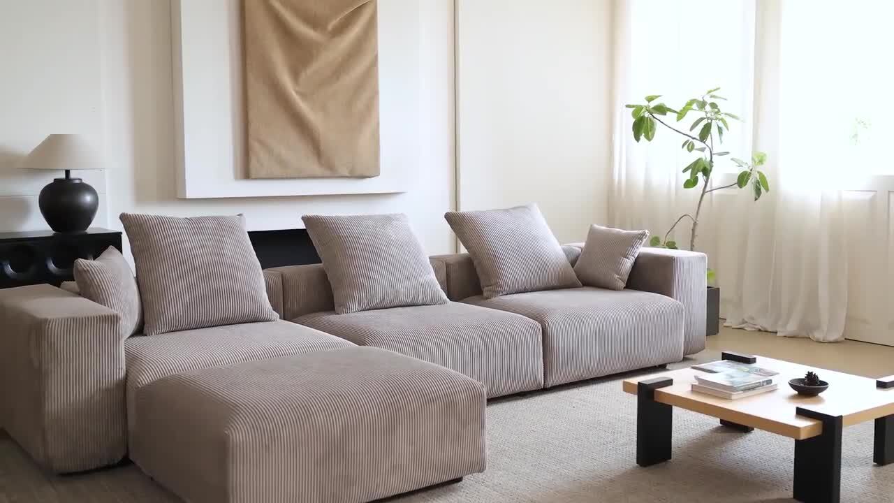 A contemporary living room featuring a sizable black 142 in. square arm, 4-piece L-shaped corduroy polyester modern sectional sofa. A wooden coffee table sits in the center, complemented by a black lamp on a side table. A potted plant is placed near the window adorned with sheer curtains, while minimalist artwork graces the wall.