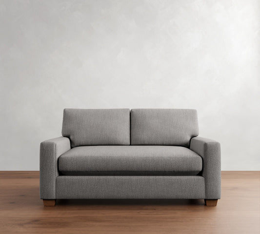 The PB Comfort Modern Square Arm Upholstered Sofa, a gray two-seater with a trendy design, is placed on a wooden floor against a textured white wall. It showcases square arms and boasts a minimalist style.