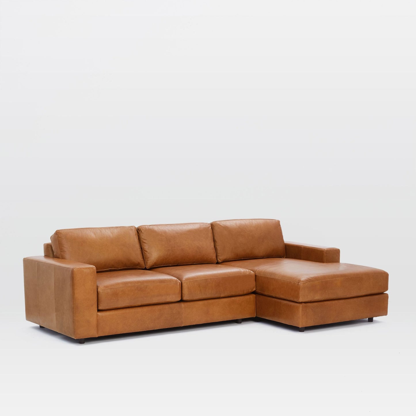 The Urban Leather 2 Piece Chaise Sectional, featuring a rich brown hue and composed of leather upholstery with three cushions and a right-side chaise lounge, is set against a simple white background.