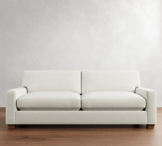 The PB Comfort Modern Square Arm Upholstered Sofa, adorned with two cushions, is set against a light gray wall on a wooden floor.