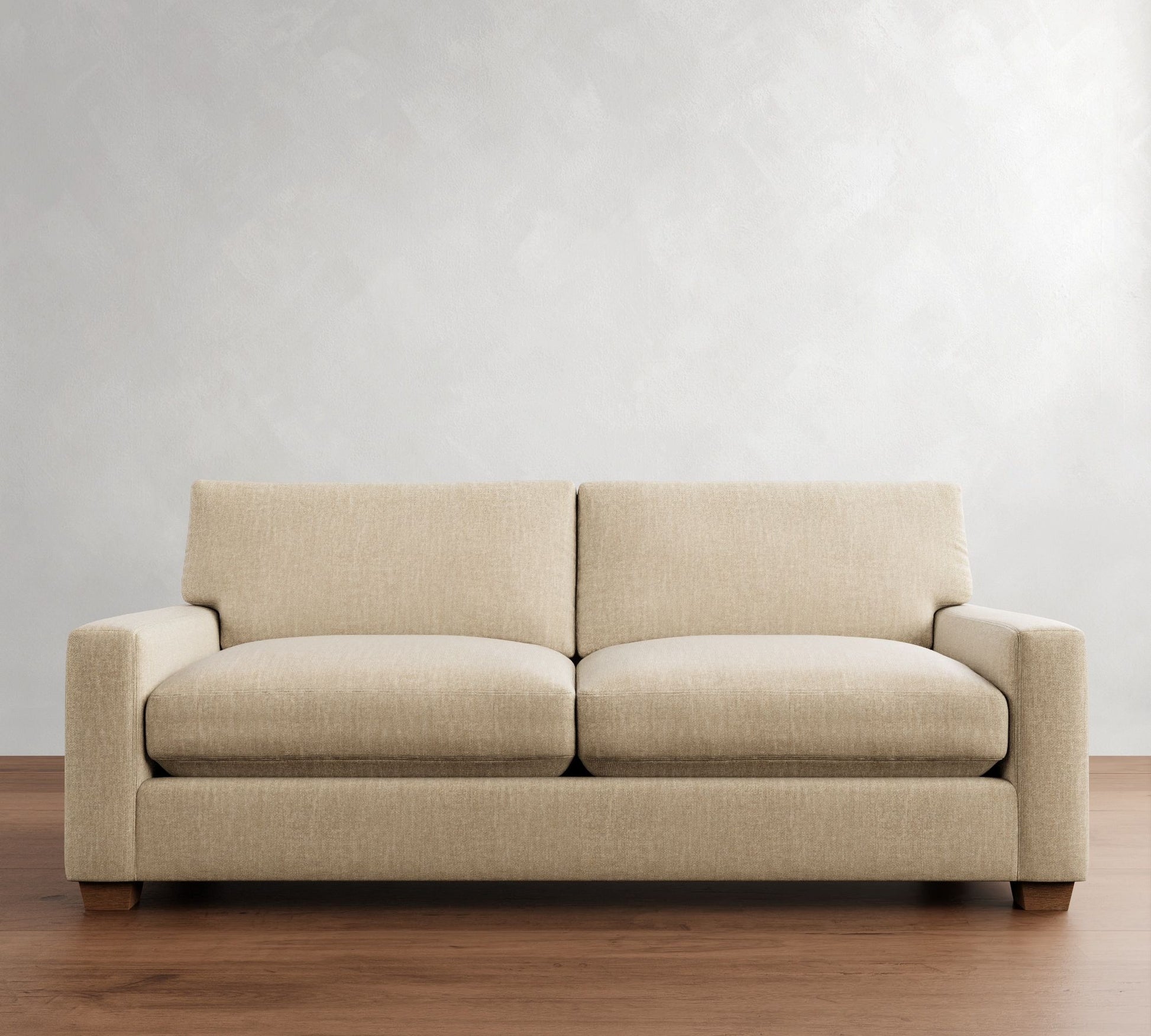 The PB Comfort Modern Square Arm Upholstered Sofa, featuring a simple and modern design with square armrests and wooden legs, is positioned against a light gray wall with its beige fabric complementing the wooden floor. It comes with two seat cushions and two back cushions.