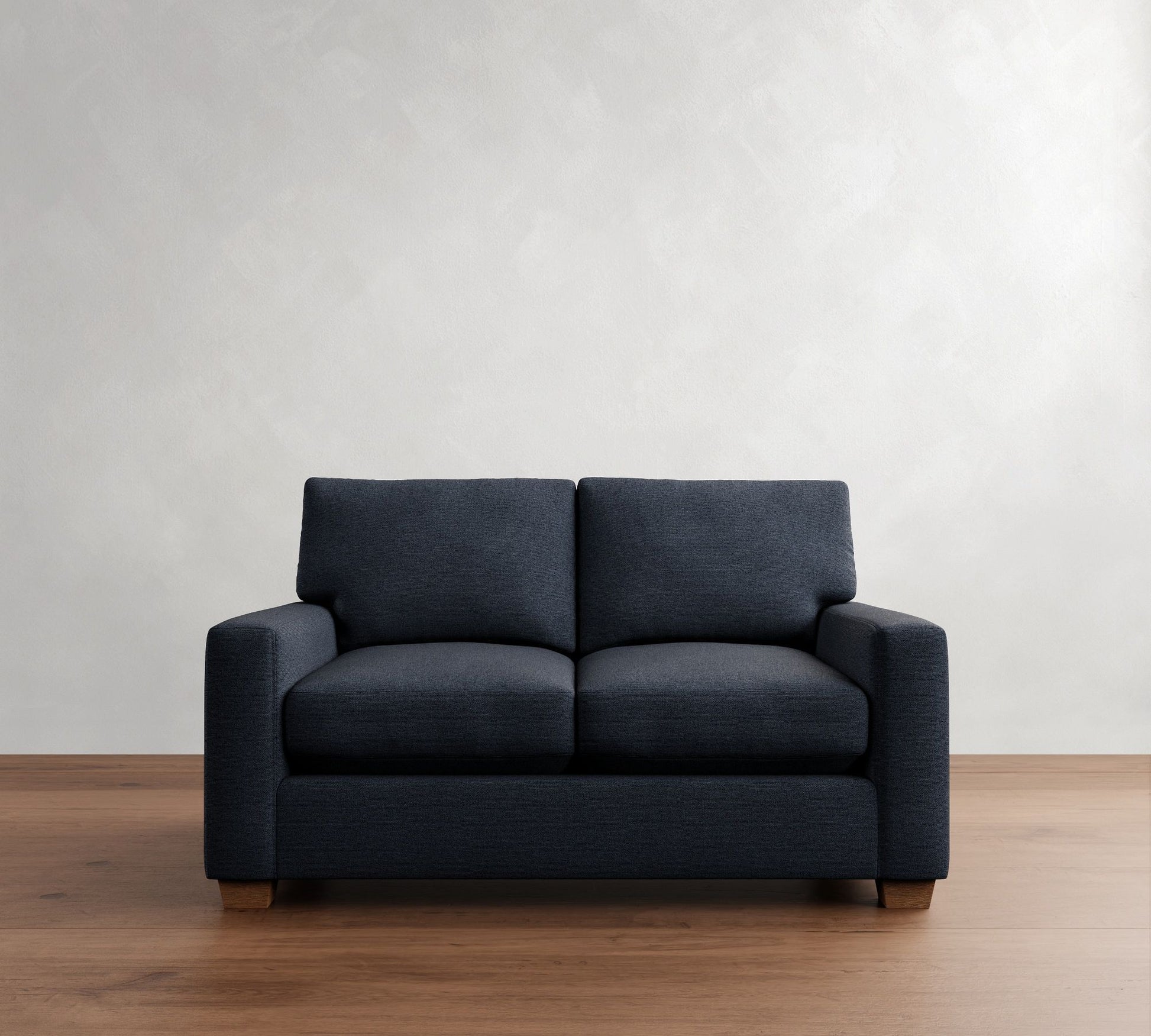 The PB Comfort Modern Square Arm Upholstered Sofa, featuring a two-seater design in dark grey with a minimalist style, sits on a wooden floor against a plain white wall.