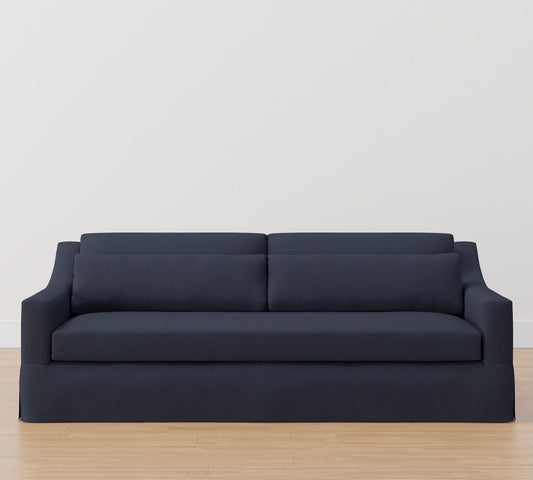 The York Slope Arm Deep Slipcovered Sofa in dark gray rests on a light wooden floor against a plain white wall. With its two large back cushions and sleek, modern design, it offers a minimalist aesthetic.