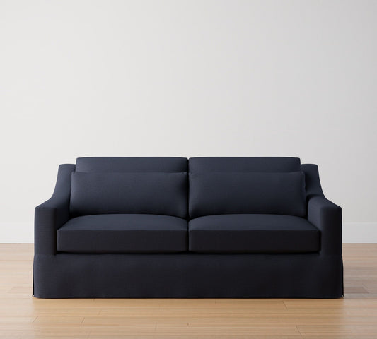 The York Slope Arm Deep Slipcovered Sofa in dark blue, featuring sleek cushions and accommodating two seats, rests on a light wooden floor against a plain white wall. Its straight lines and minimalist design offer a modern and clean aesthetic to the room.