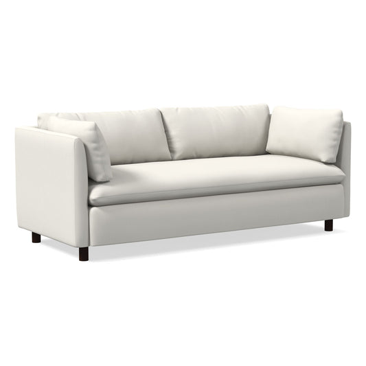 The Shelter Sleeper Sofa is a chic, modern piece with a minimalist white design. It boasts plush cushions and straight armrests, all supported by short, dark wooden legs, making it perfect for a contemporary living space.