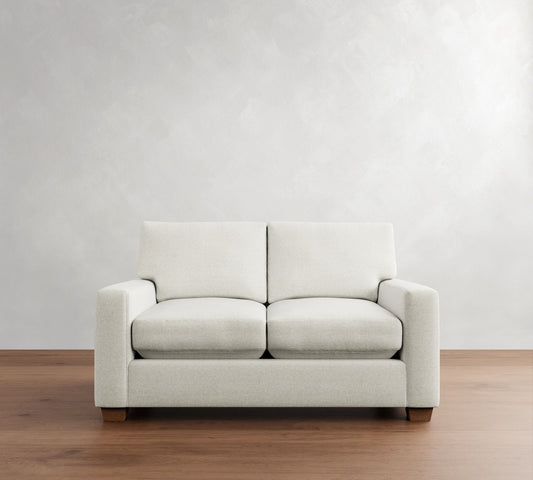 The PB Comfort Modern Square Arm Upholstered Sofa, featuring a minimalist design and light gray upholstery, is placed on a wooden floor against a plain, light gray wall. The sofa includes two cushions and stands on sturdy wooden legs.