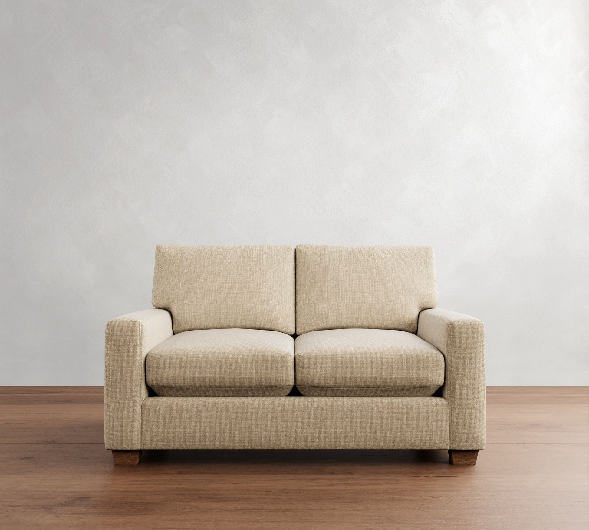 A beige PB Comfort Modern Square Arm Upholstered Sofa with wooden legs is placed on a wooden floor against a neutral textured wall. This minimalist two-seater sofa includes square arms, two seat cushions, and two back cushions.