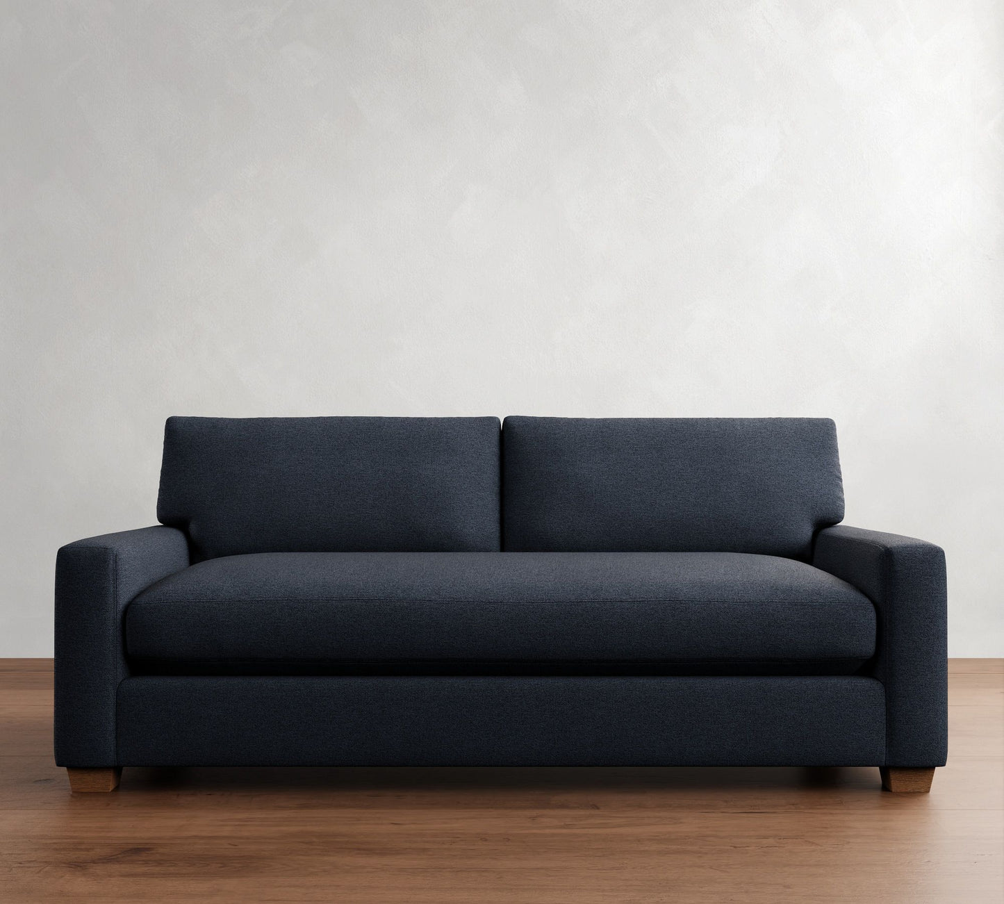 The PB Comfort Modern Square Arm Upholstered Sofa, featuring a modern dark grey hue with plush cushions, is positioned on a wooden floor against a light grey textured wall. The sofa showcases a minimalist design with clean lines and wooden legs.