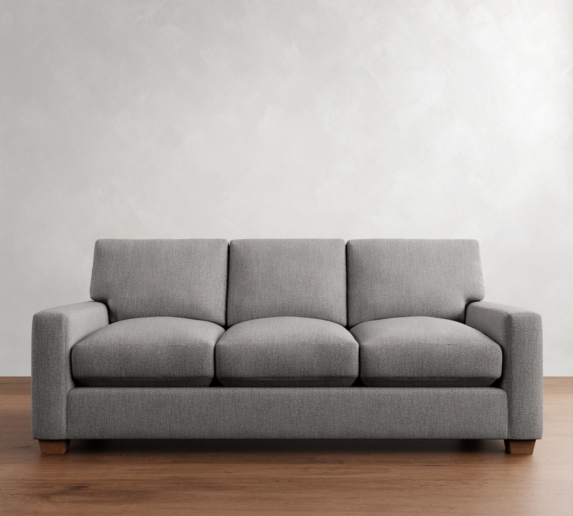 The PB Comfort Modern Square Arm Upholstered Sofa, a contemporary gray three-seater with plush cushions, is placed centrally in a minimalist space. The backdrop features a white textured wall, and the floor boasts a warm wooden finish.