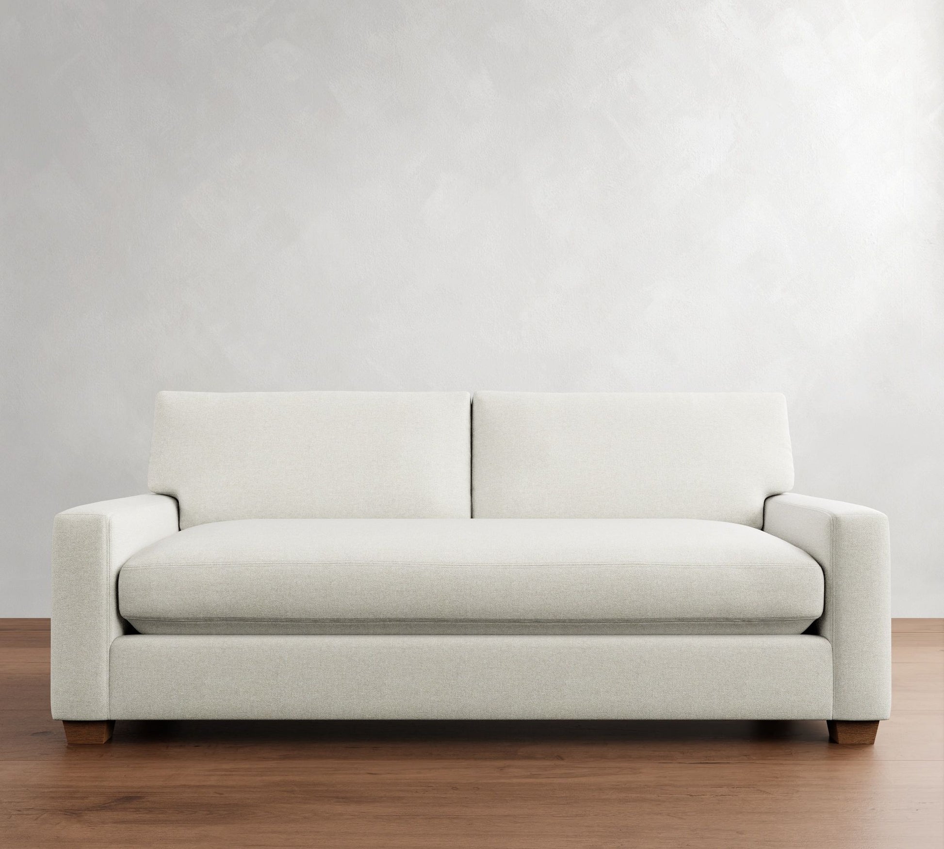 The PB Comfort Modern Square Arm Upholstered Sofa, characterized by its light gray hue and sleek design with clean lines and rectangular cushions, complements a minimalist living room setting as it rests on a wooden floor against a softly muted gray wall.