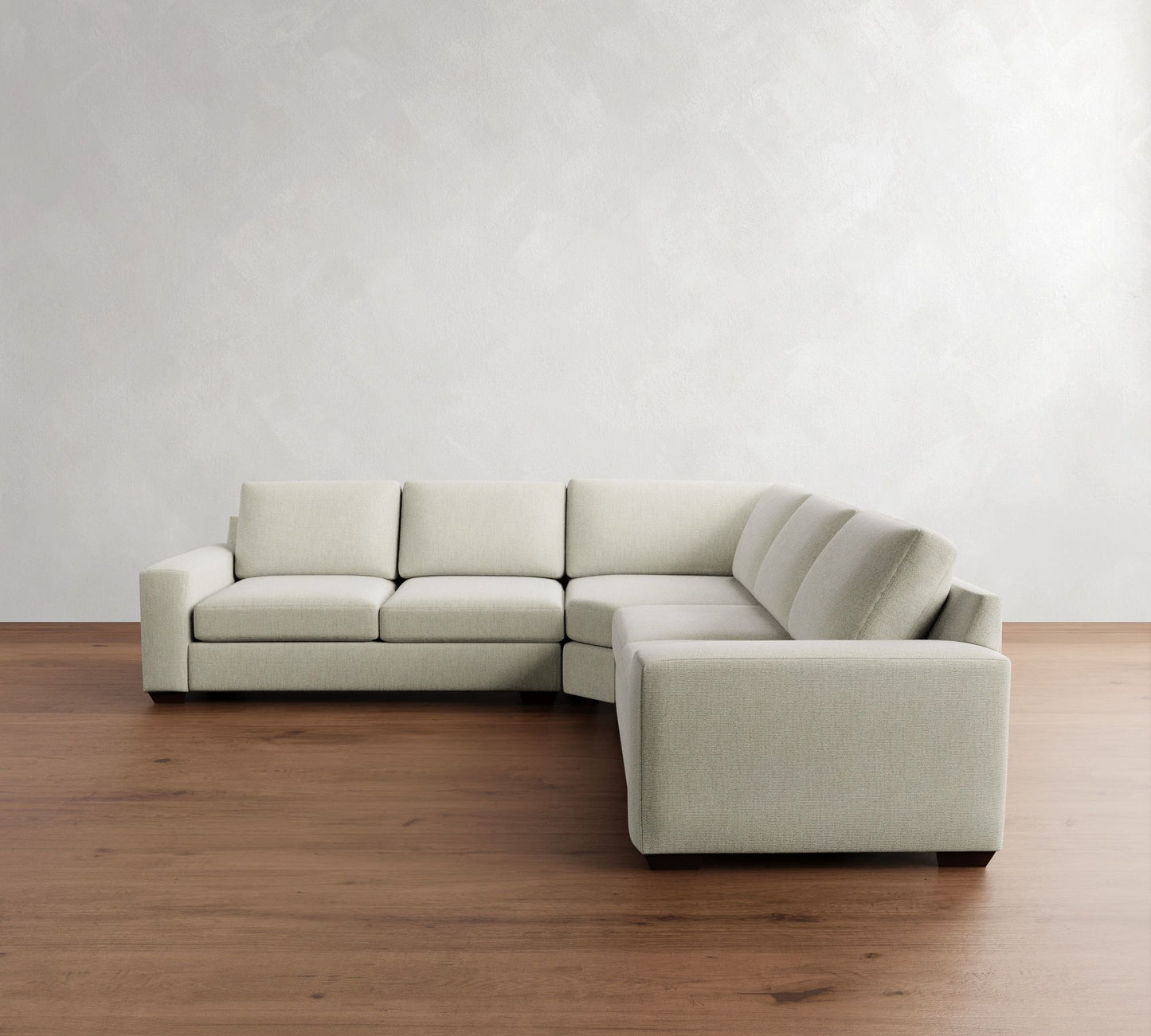 The Big Sur Square Arm Upholstered 3-Piece L-Shaped Wedge Sectional, featuring a minimalist light gray design with clean lines, is set upon a wooden floor against a textured white wall. The sectional forms an L-shape, offering a modern and spacious seating area.