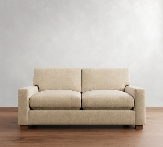 The PB Comfort Modern Square Arm Upholstered Sofa, featuring two seat cushions and two back cushions, is arranged against a light gray textured wall. It sits on a wooden floor and boasts square armrests and wooden legs.