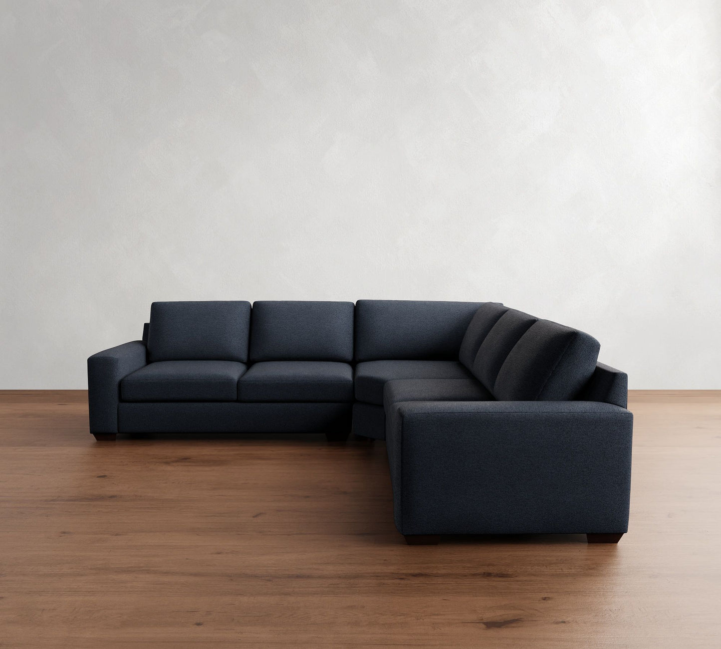 A minimalist living room showcases the Big Sur Square Arm Upholstered 3-Piece L-Shaped Wedge Sectional in dark gray, positioned on a wooden floor. The background features a plain light-colored wall, contributing to a simple and modern aesthetic.