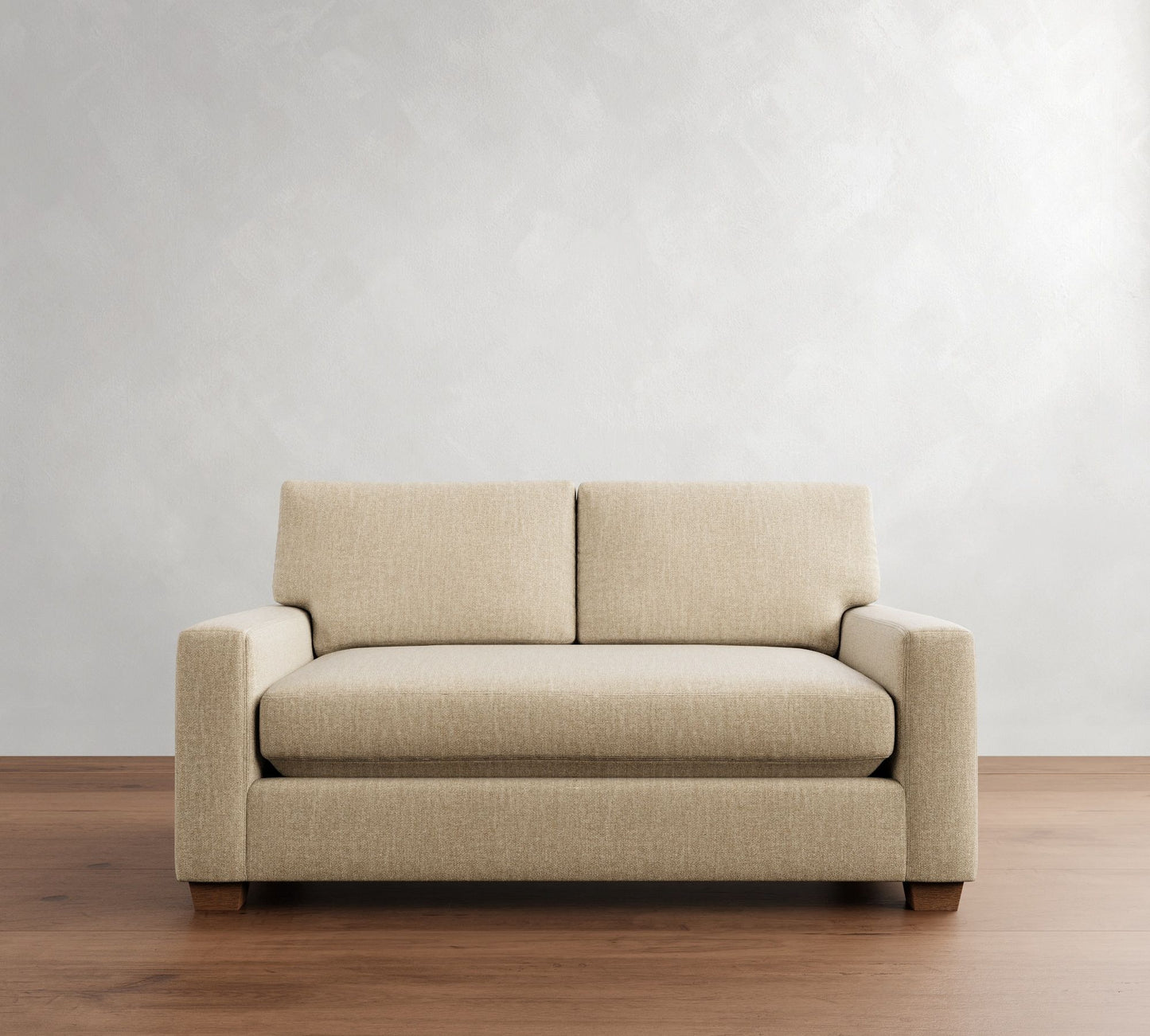 The PB Comfort Modern Square Arm Upholstered Sofa, in a minimalist beige fabric, is placed on a wooden floor against a light gray wall, creating a cozy and neutral living space.