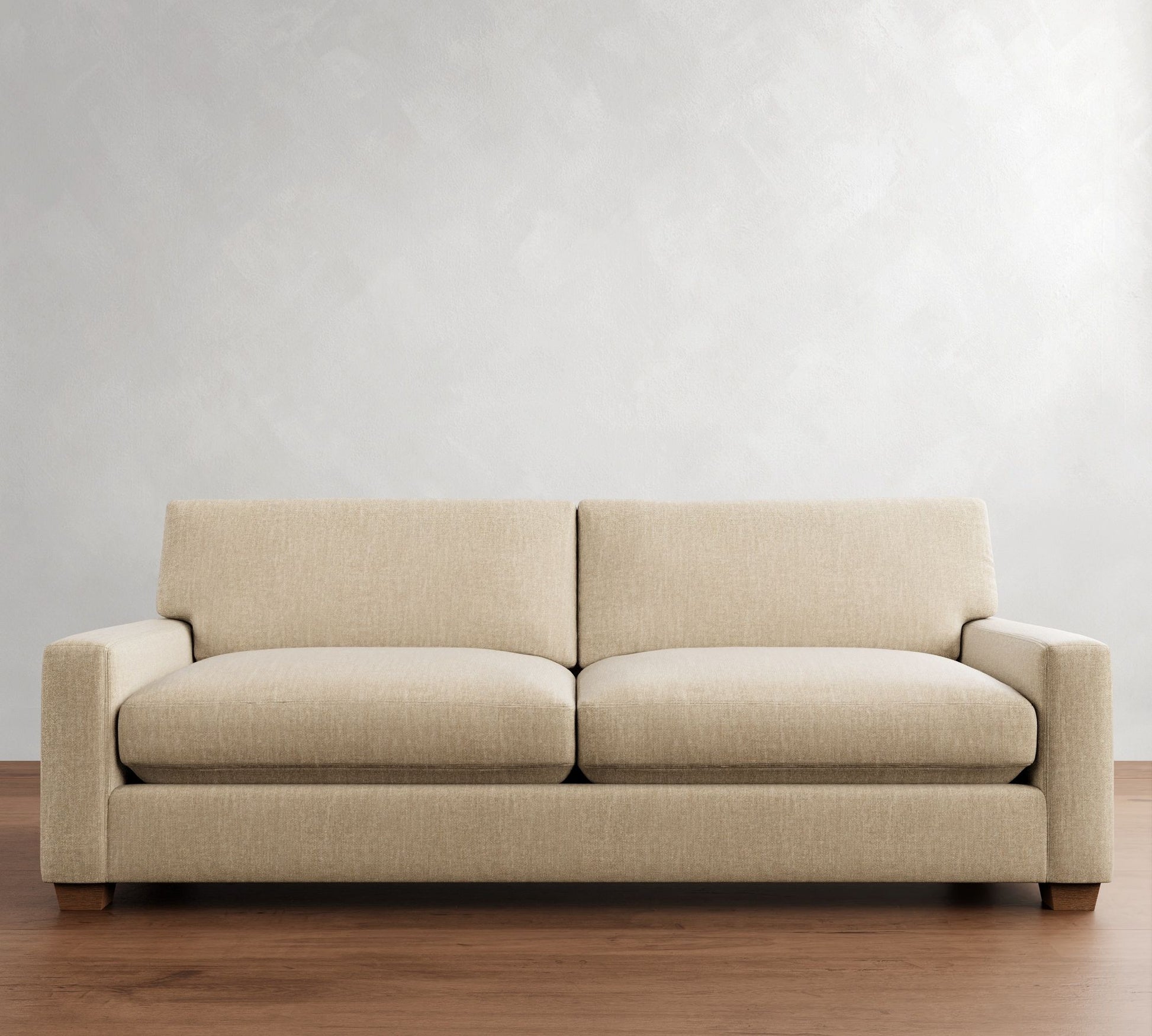 The PB Comfort Modern Square Arm Upholstered Sofa, with its modern design and beige upholstery, rests on a wooden floor against a simple light gray wall. It features two large cushions, armrests, and a sturdy base, contributing to its minimalist and elegant look.