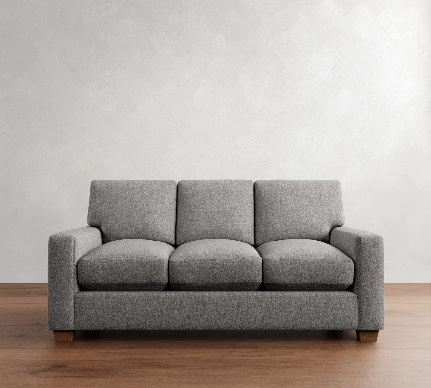 The PB Comfort Modern Square Arm Upholstered Sofa, with its modern gray upholstery and square cushions, rests on a wooden floor against a light textured wall. Its wooden legs add to the minimalist and contemporary design.