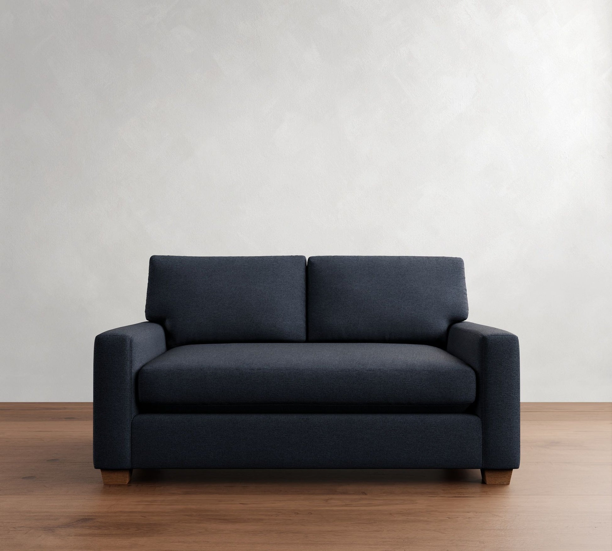 The PB Comfort Modern Square Arm Upholstered Sofa, in a dark blue hue, is positioned against a light gray textured wall on a wooden floor. Its simple and minimalist design brings an elegant and sleek appearance to the setting.