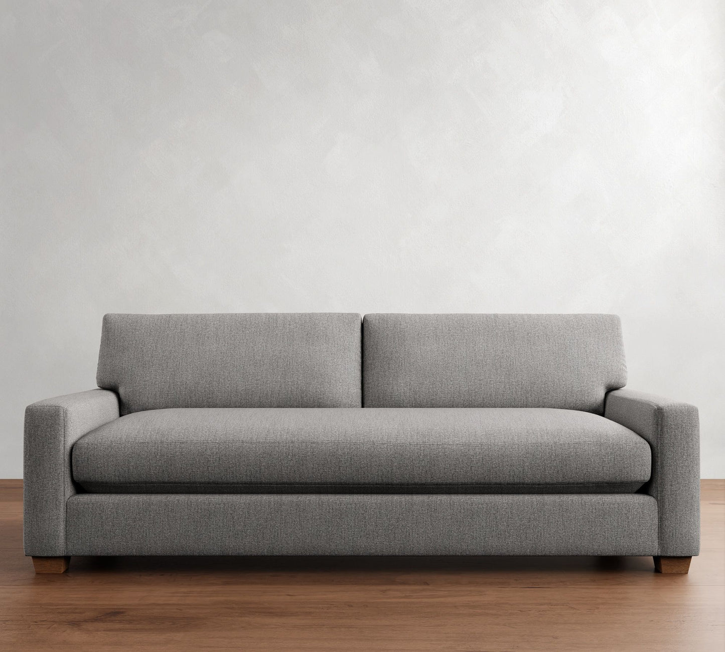 The PB Comfort Modern Square Arm Upholstered Sofa, featuring a sleek gray fabric design, rests on a wooden floor against a plain light-colored wall. This sofa comes with two seat cushions and a low backrest, offering a minimalist and modern appearance.