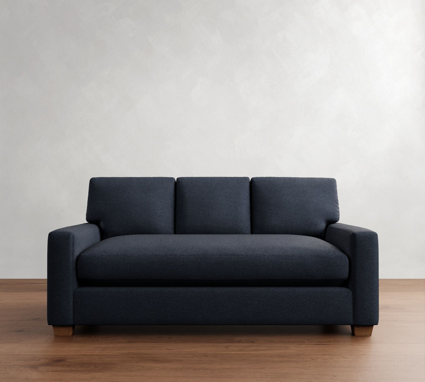 The PB Comfort Modern Square Arm Upholstered Sofa, in dark gray, is centered in a room with light-colored walls and a wooden floor. This sofa features a simple and modern design with square armrests and wooden legs, complemented by three cushions.