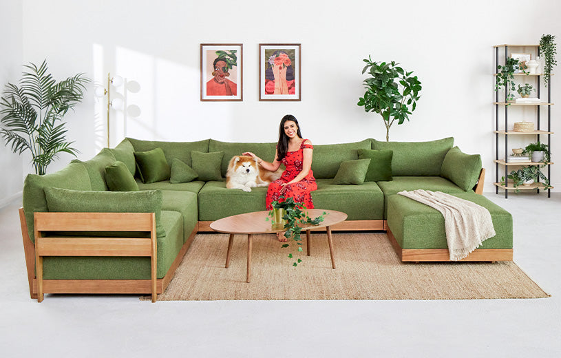 A woman in a red dress sits on a large green sectional sofa with a dog. The room is adorned with green plants, framed artworks on the wall, and a wooden coffee table with a small plant. The decor features a mix of modern and natural elements.