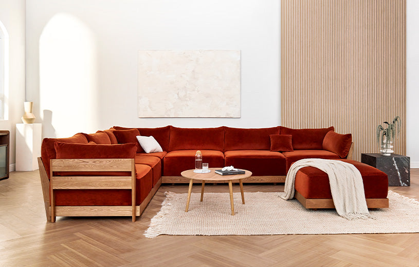 A modern living room featuring a large velvet L-shaped sectional sofa in a rich rust color. The sectional is complemented by a circular wooden coffee table and a light-colored rug on a herringbone hardwood floor. The wall behind has minimalistic decor and a large, abstract art piece.