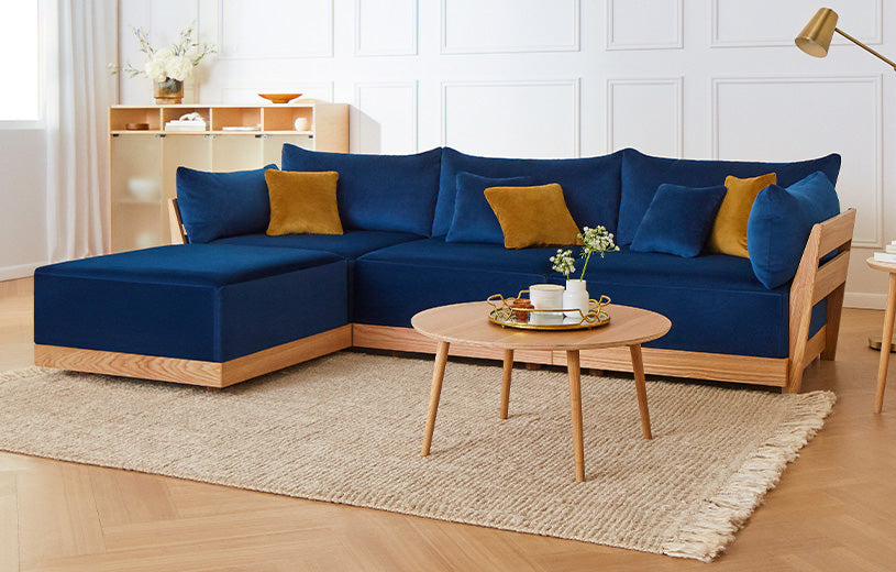 A modern living room featuring a blue sectional sofa with mustard and blue throw pillows, set on a beige textured rug. A small round wooden coffee table holds a white tray with a teapot and a small plant. The background features a minimalist wooden shelf and white paneled walls.