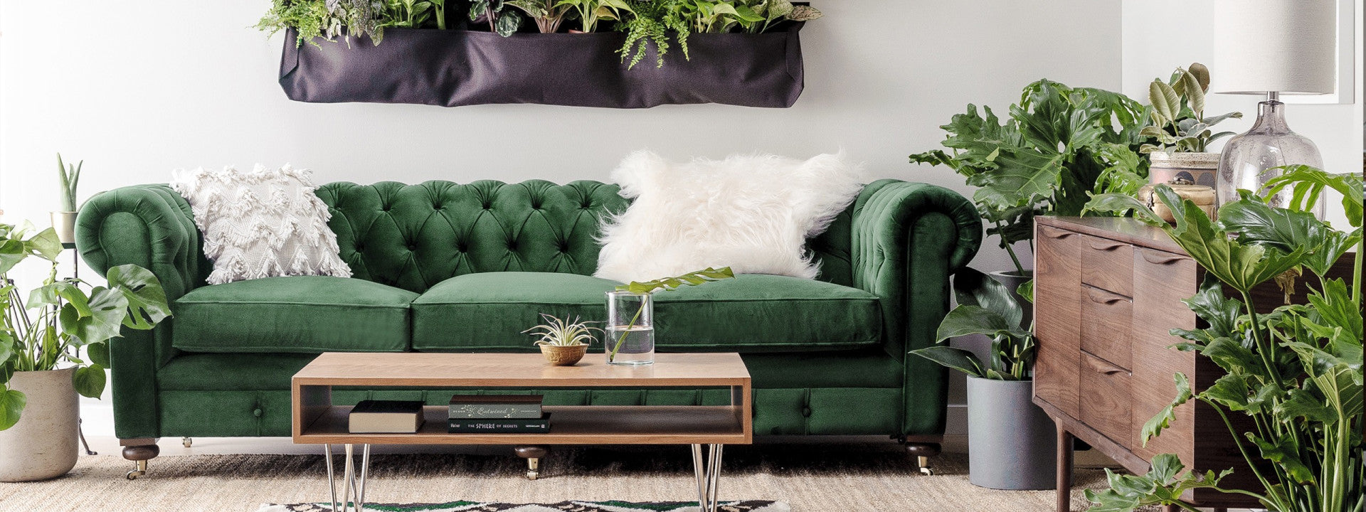 A stylish living room features a green velvet sofa with decorative pillows, surrounded by numerous green plants. Above the sofa is a vertical garden with lush greenery. A wooden coffee table with books and a glass of water stands in front of the sofa, and a wooden dresser is on the right.