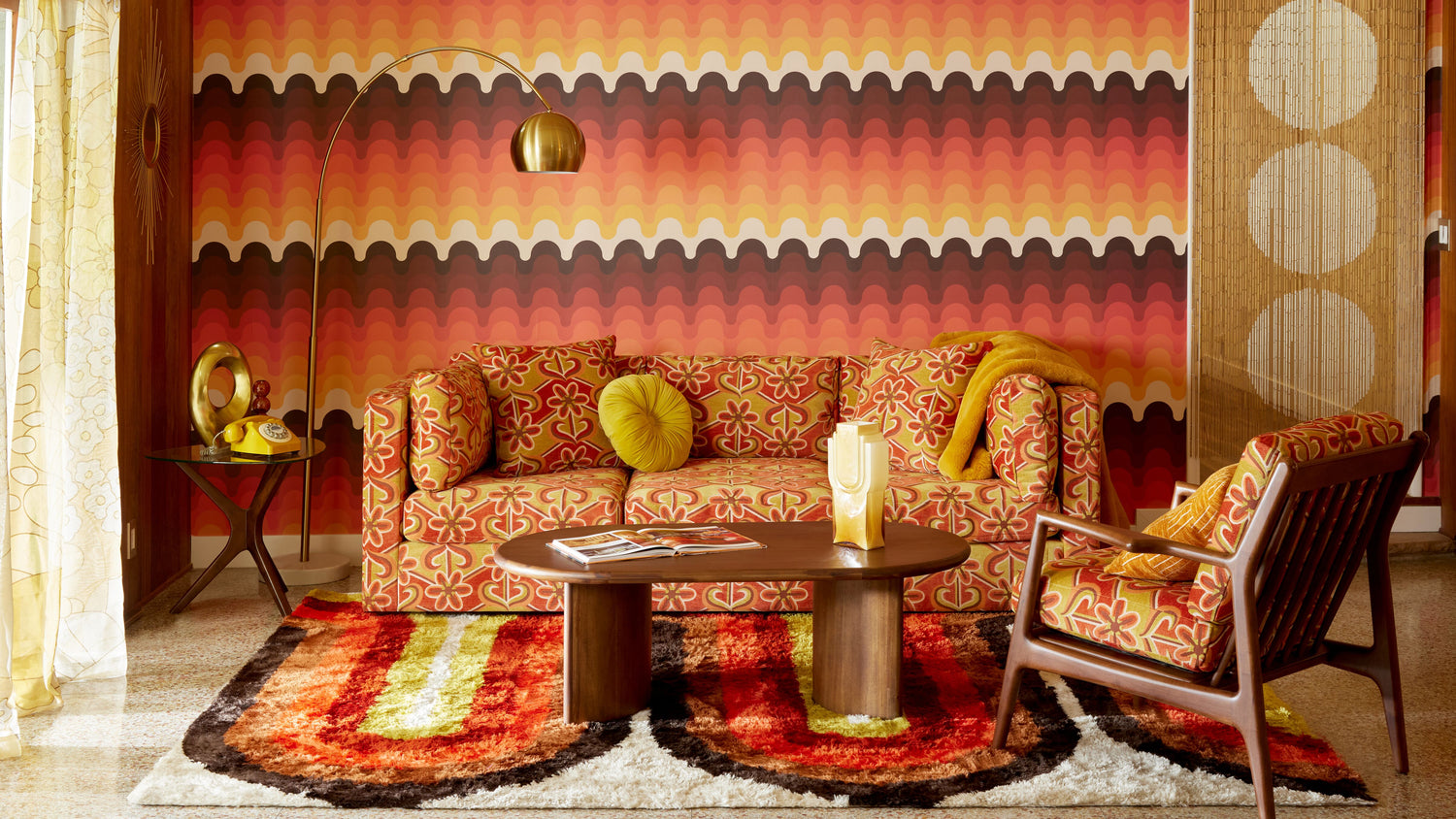 A retro living room features vibrant orange, red, and yellow patterns. The room includes a floral-patterned sofa with green cushions, a matching armchair, a wooden coffee table with magazines, a tall floor lamp, and a retro wall and carpet design.