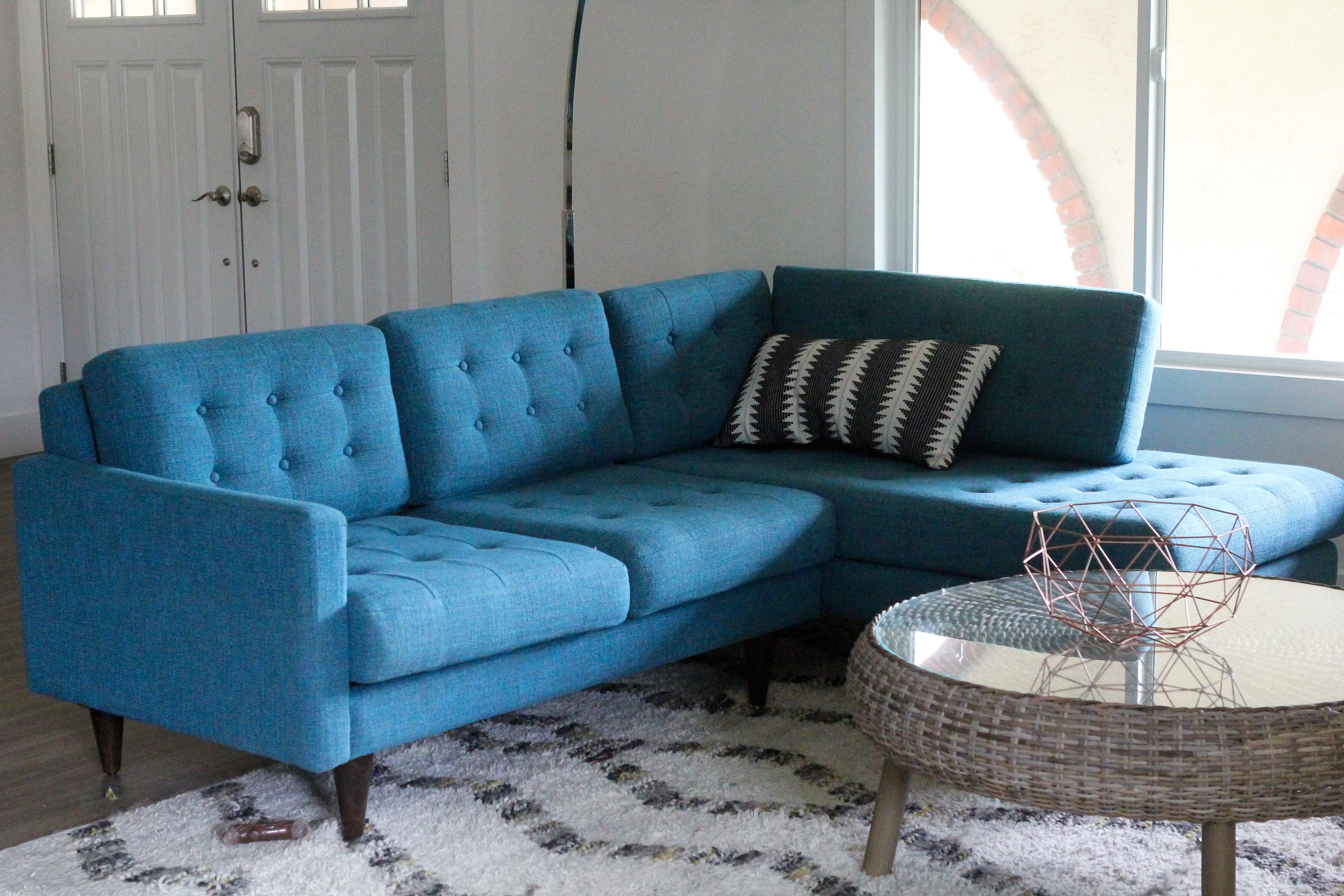 A modern living room featuring a blue, tufted sectional sofa with an angled backrest. A decorative pillow with a geometric pattern is placed on the sofa. A round wicker coffee table with a geometric wire decor item is on a light and dark patterned rug.