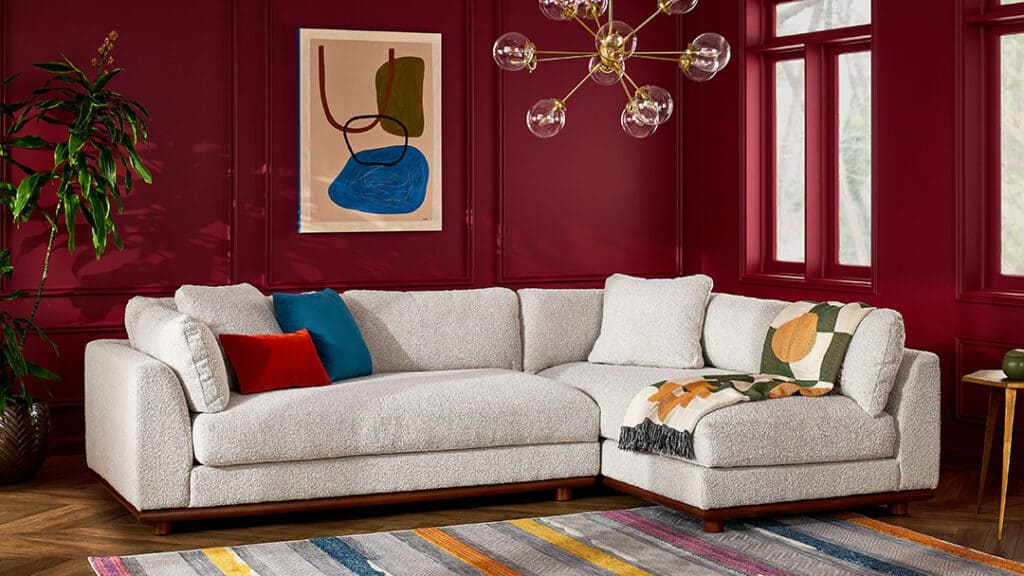 A modern living room with deep red walls featuring a white sectional sofa adorned with colorful cushions and a patterned throw. A contemporary chandelier with globe lights hangs above. The space is accented by a large abstract painting and a striped rug.