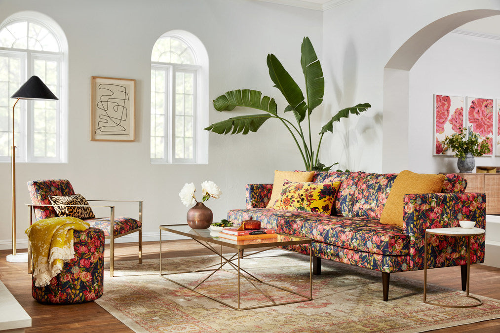 A stylish living room with a floral-patterned sofa and armchair, adorned with yellow and red cushions. A glass coffee table holds books and a floral arrangement. Large green plant and framed art add to the decor. Natural light streams through arched windows.